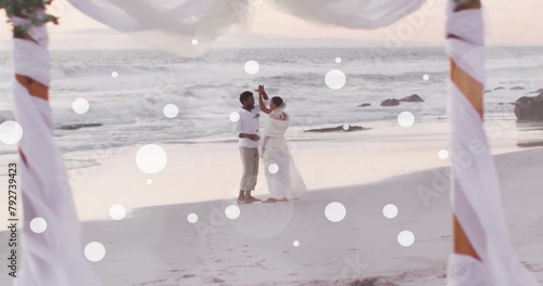 Image of white spots over african american bride and groom dancing on beach at wedding