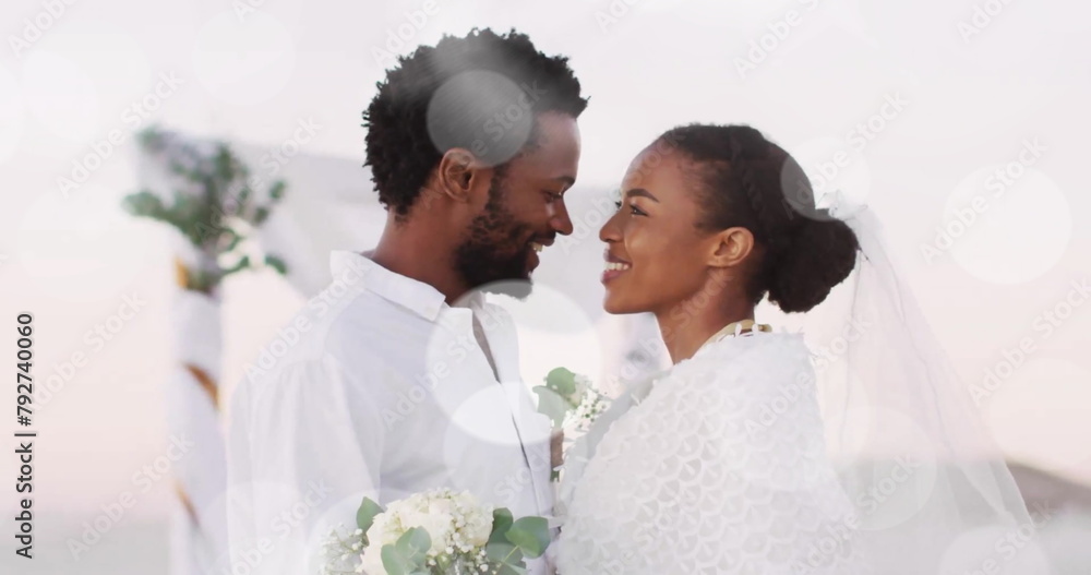 Naklejka premium Image of light spots over happy african american bride and groom embracing on beach at wedding