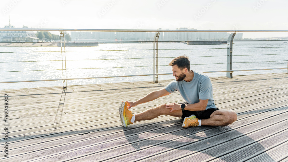 A man stretches his leg on a sun-kissed waterfront boardwalk, the glistening river complementing his morning fitness routine.