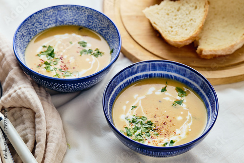 Two bowls of creamy potato soup, spoons, napkin and wooden cutting board with a loaf of bread and cut pieces. The surface of the soup is decorated with cream, croutons, parsley and ground red pepper. photo