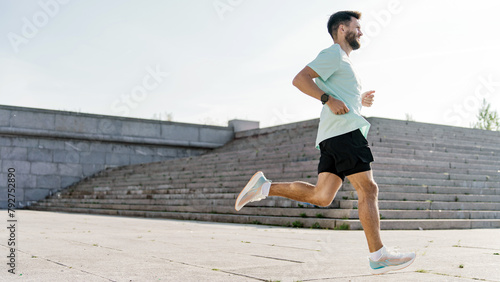 A bearded runner with a focused gaze propels forward in a dynamic stride along a vast urban staircase, under the open sky. © muse studio
