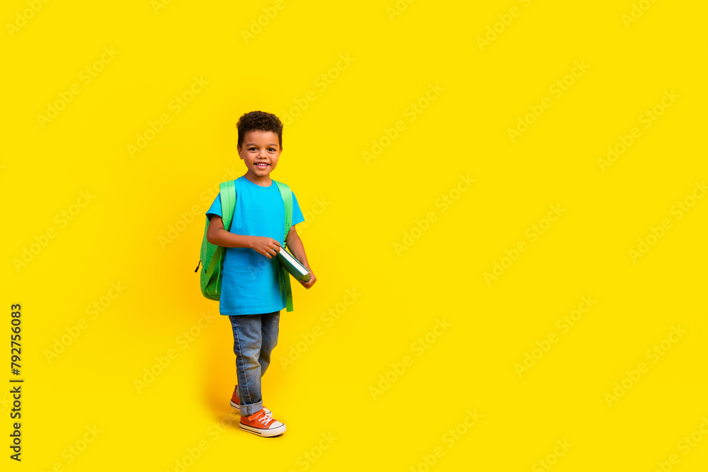 Full size photo of diligent little boy wear casual blue t-shirt rucksack holding book go on lessons isolated on vivid yellow background Full size photo of diligent little boy wear casual blue t-shirt rucksack holding book go on lessons isolated on vivid yellow background