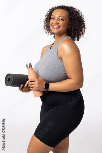 Biracial plus size model with yoga mat and water bottle, smiling, white background