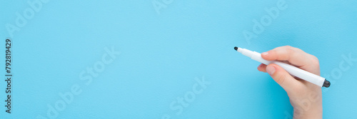Baby boy hand holding black white color pen and writing on light blue table background. Pastel color. Closeup. Wide banner. Empty place for text. Top down view.