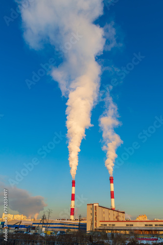 Two tall red and white chimneys spewing smoke into the sky. The sky is clear and blue