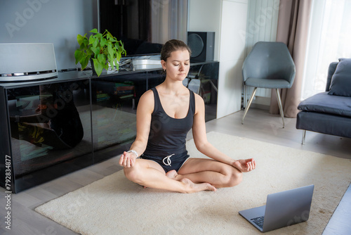 Young woman in sportswear doing yoga exercises on mat in living room. Online workout, fitness, practicing and meditation at home