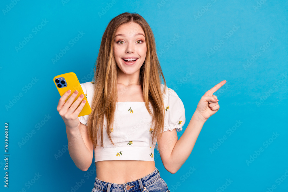 Photo of excited girl hold cell gadget point demonstrate empty space isolated blue color background Photo of excited girl hold cell gadget point demonstrate empty space isolated blue color background