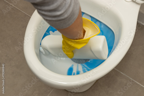 Close up of hand wearing a yellow protective glove and applying a blue liquid toilet cleaner to clean and disinfect a modern, white, ceramic toilet bowl