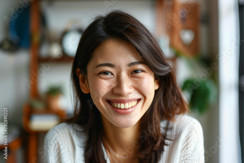 A joyful 20s Japanese girl poses for a headshot at home. The happy young woman looks directly at the camera, showcasing her white teeth in a warm smile while laughing and chatting on a video call.