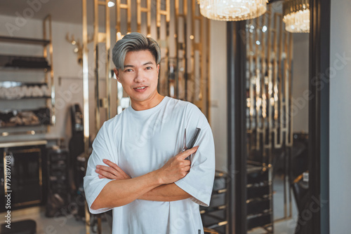 Happy asian professional Hairdresser or hair stylist man standing confidence with arm crossed with smile and holding hairdressing equipment while looking at camera in beauty salon and barber shop © oatawa