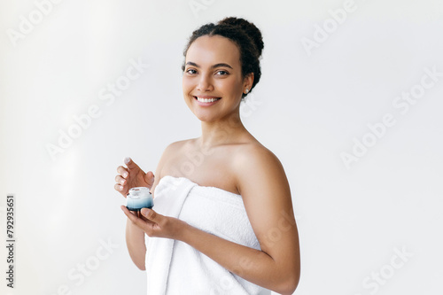 Beauty concept. Skincare. Gorgeous hispanic or brazilian young woman, wrapped in white towel, holding nourishing body cream in a hand, preparing to apply to skin after shower, looks at camera, smiling