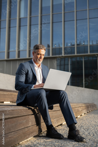 Focused mature successful businessman working, typing on laptop sitting outdoors at office building. Handsome older man entrepreneur in suit using wireless pc computer for online business. Vertical