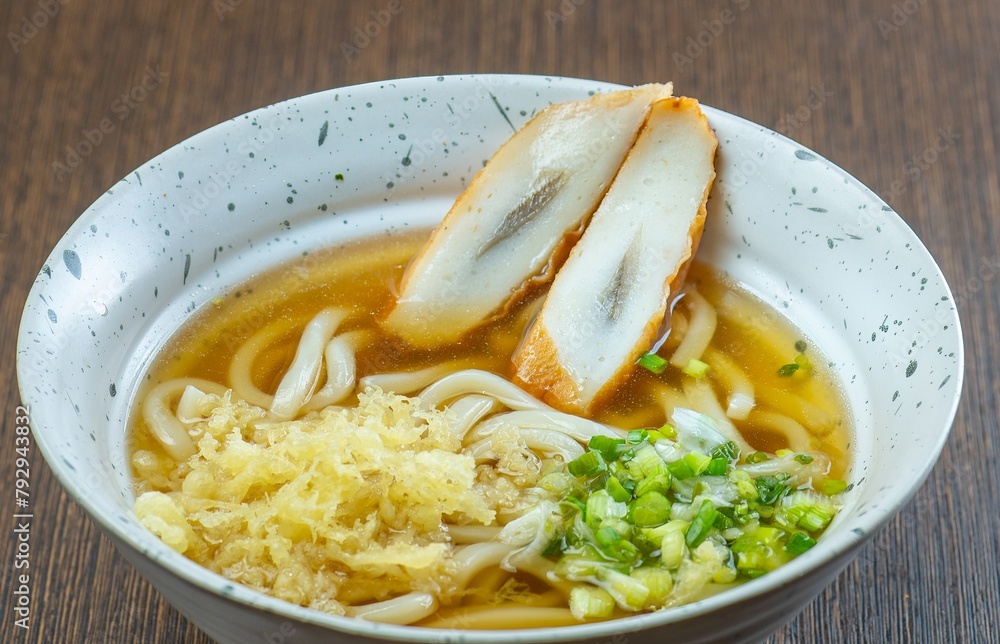 A bowl of udon noodles with a clear broth, topped with tempura and green onions. A bowl of udon noodles with a clear broth, topped with tempura and green onions.