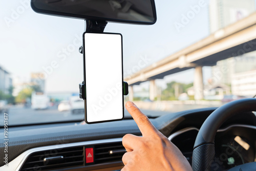 Close-up smartphone with white mockup on screen, background of car steering wheel. A person is pointing at a phone mounted on the dashboard of a car.
