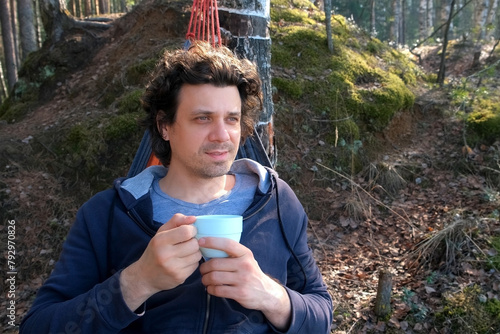 Portrait curly man relaxing lying in hammock drinking tea in forest on nature. Resting relaxed brunet male enjoying time in camping in woodland. Tourist, traveler, camper contemplating outdoors.