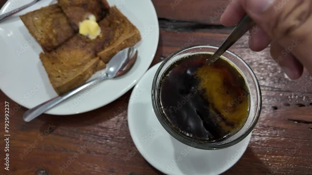 Someone is stirring a cup of hot butter coffee with spoon and eating toast for breakfast in Kopi Tiam. Someone is stirring a cup of hot butter coffee with spoon and eating toast for breakfast in Kopi Tiam.
