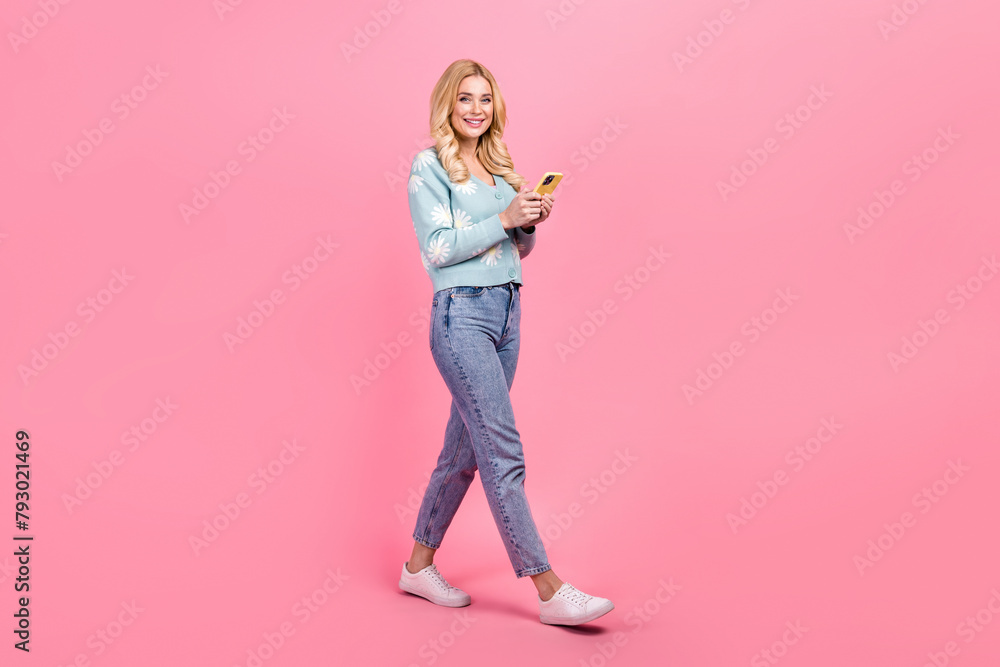 Full size photo of lovely young woman hold device walking dressed stylish flower print garment isolated on pink color background Full size photo of lovely young woman hold device walking dressed stylish flower print garment isolated on pink color background