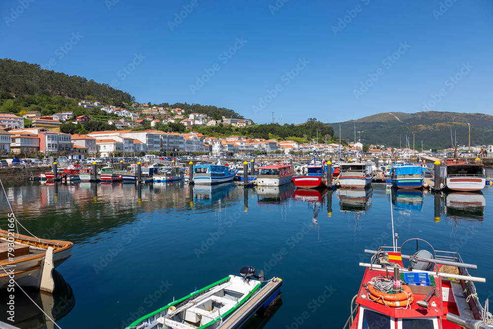 A serene marina of Muros in Spain with various boats docked in calm waters, with quaint hillside houses and clear blue sky in the background - obrazy, fototapety, plakaty 