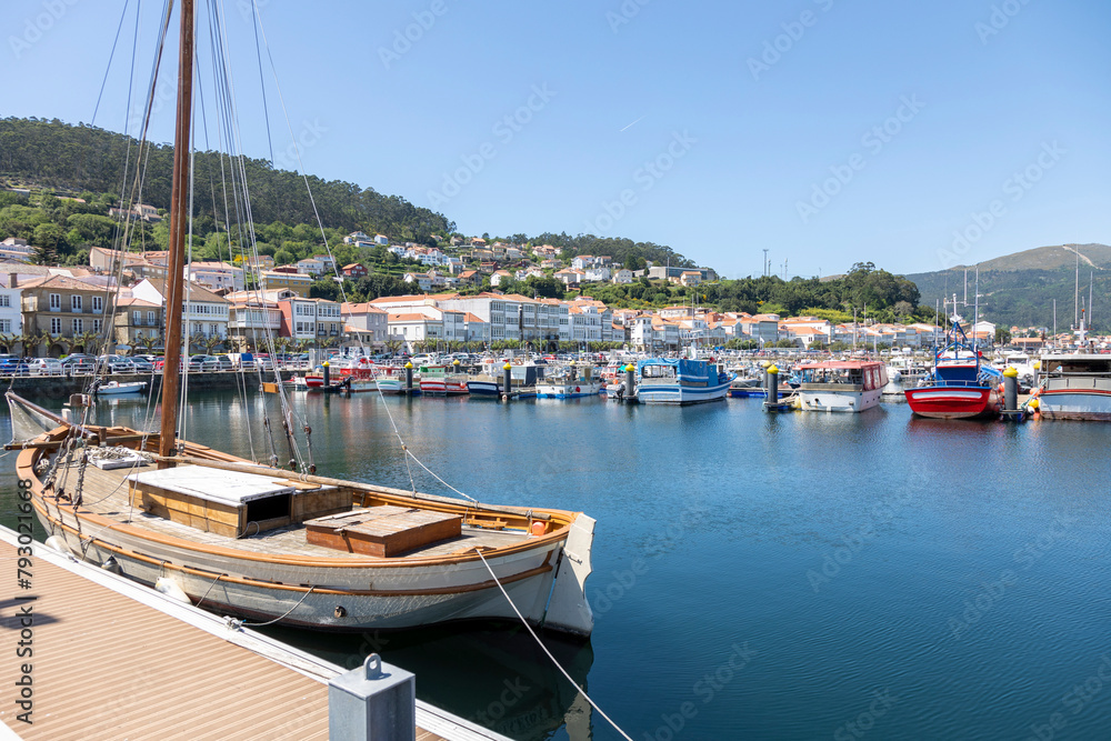 A serene marina of Muros in Spain with various boats docked in calm waters, with quaint hillside houses and clear blue sky in the background - obrazy, fototapety, plakaty 
