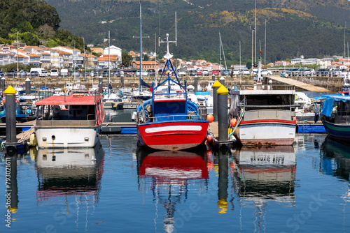 A serene marina of Muros in Spain with various boats docked in calm waters, with quaint hillside houses and clear blue sky in the background