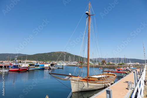 A serene marina of Muros in Spain with various boats docked in calm waters, with quaint hillside houses and clear blue sky in the background