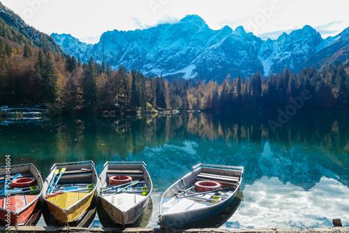 Colorful wooden boats on Fusine Lake (Laghi di Fusine) with scenic view of snow capped Julian mountain range in Tarvisio, Friuli-Venezia Giulia, Italy, Europe. Water reflection in green alpine lake