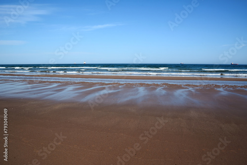A wide beach with shallow smooth sand. Several ships are sailing in the sea in the distance