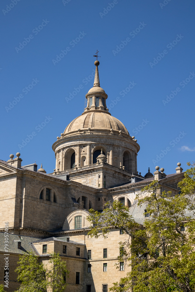 The sculptural details and the emblematic dome of the Monastery of El Escorial with roofs and gardens with trees.