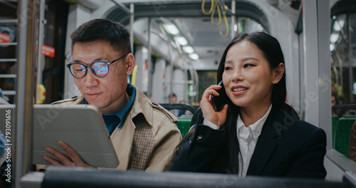 Asian man with glasses and raincoat focused in his tablet device. Young girl smiling while talking with someone. Beginning to look around. Worried about missing correct train stop. People on way home. © ihorvsn