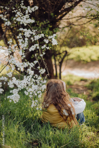 a little cute girl with long hair sits on the grass, enjoys the spring, admires the flowers on a blooming tree on a sunny day. smiling, enjoying the good weather. walk, family leisure, no face