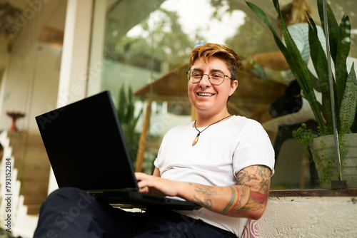 Transgender professional works on laptop, smiles in modern coworking space. Inclusive workplace, creative freelancing, comfy setting. Tattooed, gender diversity, productive remote working.