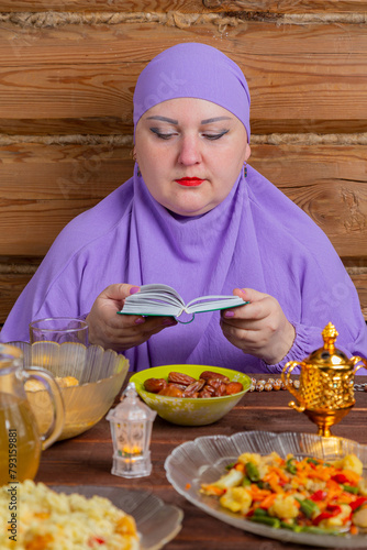 A Muslim woman in a lilac hijab at the table reads a dua from a book, a prayer for the end of Ramadan, the holiday of Eid al-Fitr.