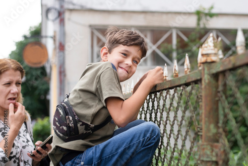 A young boy with a funny face climbed the fence in city center, bullies, his mother stands by and does not know what to do, the concept of raising children with hyperactivity