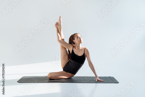 Graceful young woman pulls her leg up practicing stretching in a well-lit bright studio