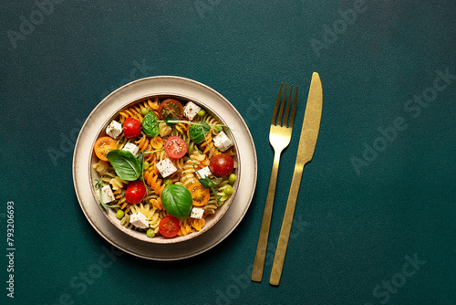 Colorful full grain fusilli pasta warm salad with feta cheese, cherry tomatoes, herbs, green pea and basil leaves on dark green background top view, copy space for your design.