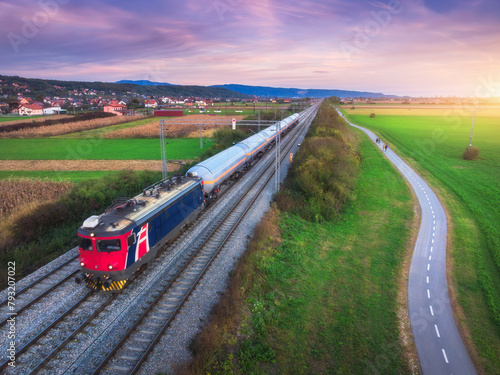 Aerial view of freight train moving near alpine village, mountains and green fields, bike path at sunset in spring. Top view of train, railroad, road, meadow in summer. Railway station in Croatia