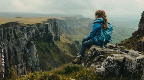 A side view of an anonymous woman in a blue jacket and boots sitting on the edge of a rocky cliff, observing the scenic view of a green mountainous valley. © Elchin Abilov