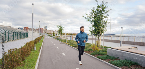 A content man jogs along a riverside pathway under cloudy skies  cityscape in the backdrop.