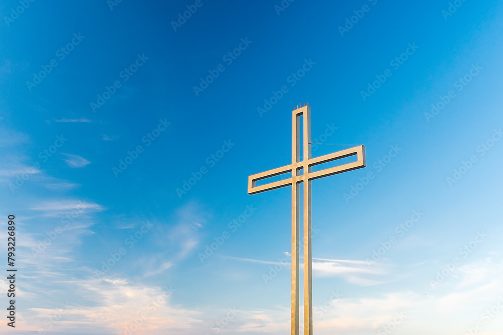 Golden cross against a background of blue sky with clouds. A minimalistic view of a gold-colored cross against the sky. Golden cross against a background of blue sky with clouds. A minimalistic view of a gold-colored cross against the sky.