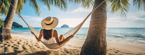 Woman Relaxing in a Hammock Between Palm Trees on a Beach. A person with sun hat lies in a hammock tied between two palm trees on a tropical sandy beach.