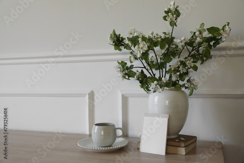 Springtime breakfast still life. Empty greeting card, invitation mockup. White ceramic vase with blooming apple tree branches. Cup of coffee, tea on wooden table, old books. Scandi home interior