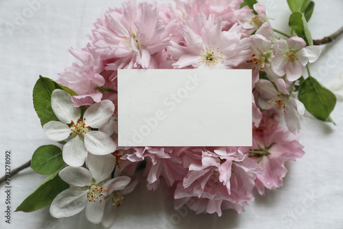 Closeup of blank business card, invitation mockup. Feminine scene with blurred pink blossoming Japanese cherry, apple tree branch. White linen table background. Flat lay, top view. Easter banner.