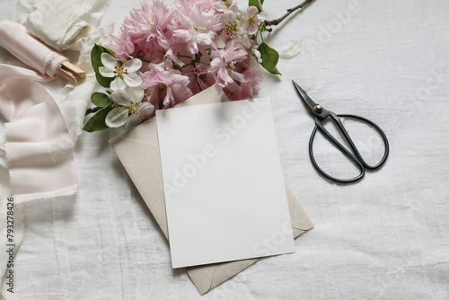 Wedding spring styled photo. Feminine scene mockup with pink blossoming Japanese cherry, apple tree branch. Blank paper greeting card, black scissors, white linen table background. Flat lay, top view.