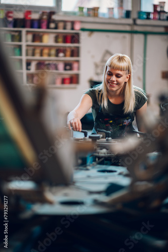 Smiling female graphic worker adjusting screen printing press at factory