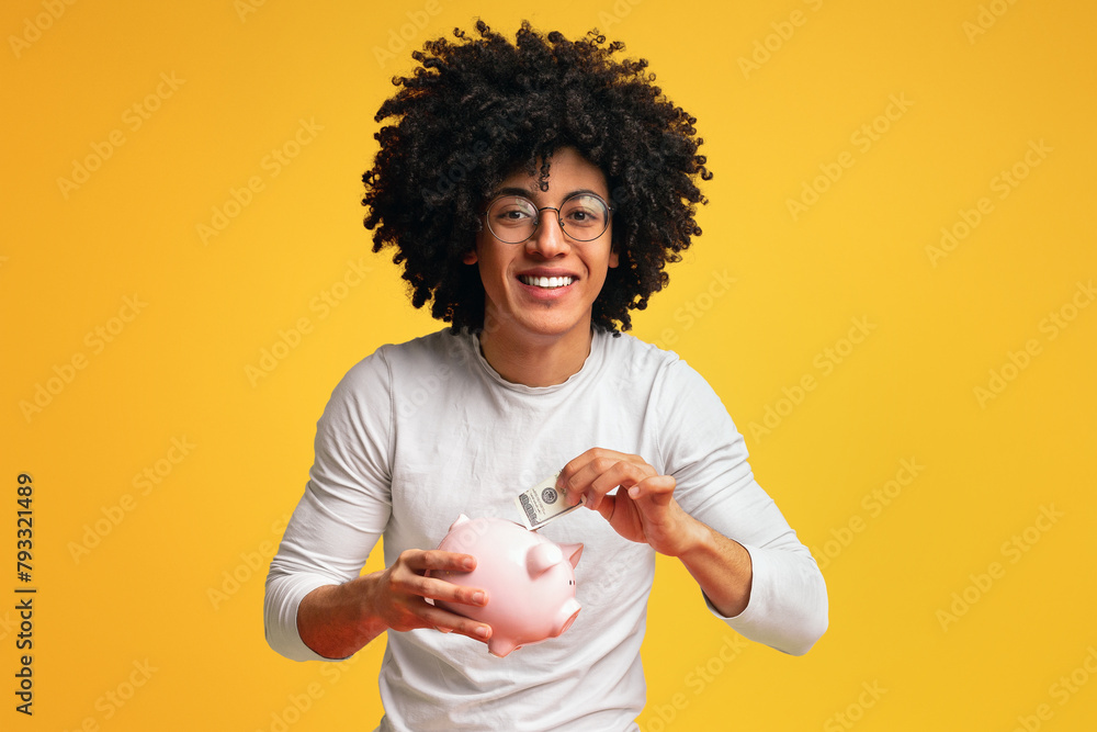 Smiling african-american man putting money in his piggy bank Smiling african-american man putting money in his piggy bank
