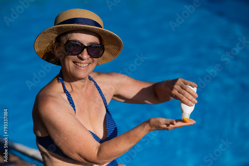 Portrait of an old woman in a straw hat, sunglasses and a swimsuit applying sunscreen to her skin while relaxing by the pool.  © Михаил Решетников