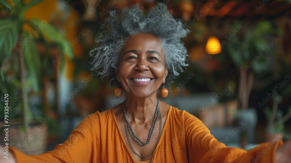 A senior woman with gray hair smiling warmly at the camera, sitting indoors with a vibrant orange attire.