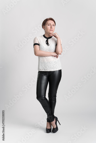 Confident businesswoman looking at camera with one hand raised to chin, standing legs crossed at ankle on white background. Female business person wearing blouse, leather trousers, high heeled shoes
