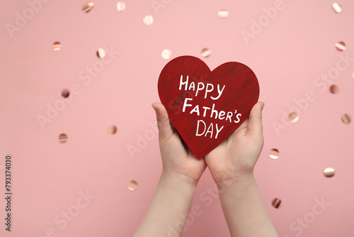 wooden red heart with white inscription happy Father's day in hands of child on pink celebration background