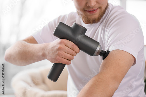 Sporty young man massaging his shoulder with percussive massager at home, closeup